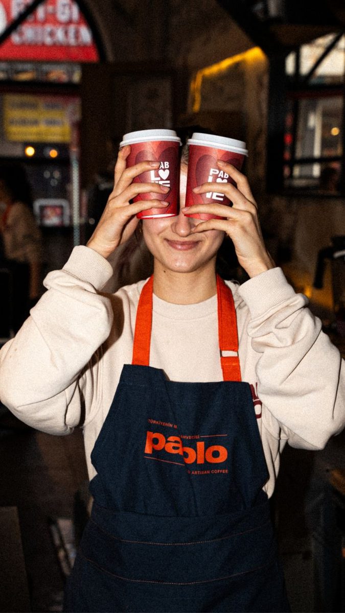 A barista in an apron holding two coffee cups over her eyes